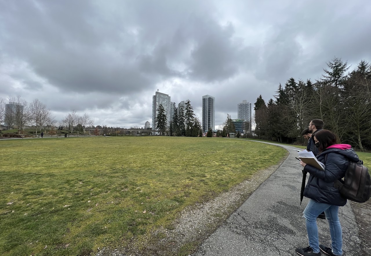 site visit photograph of Holland Park in Surrey BC showing the real-world location of the pavilion. A green field is shown with tall trees and apartment buildings in the distance.