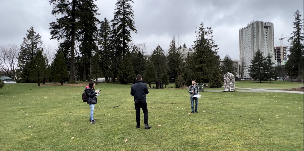 site visit photograph of Holland Park in Surrey. A green field is shown with tall trees and apartment buildings in the distance. Designers are gathered in a small group to view the location and brainstorm ideas.